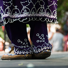 Photographie couleur, en vue rapprochée, des pieds d'une femme portant une tenue traditionnelle mauve, décorée de perles blanches.