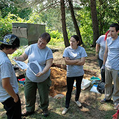 Colour photograph of trainee archaeologists, standing and looking at the illustration of a French coin held by the archaeologist.