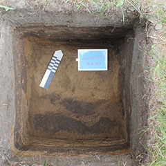 Colour photograph of a squared excavation hole at the bottom of which blackened soil can be seen.