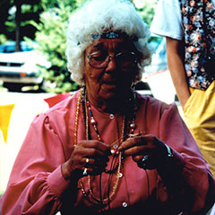 Colour photograph of a woman with short curly hair, wearing a pink shirt and several necklaces made of colored beads.